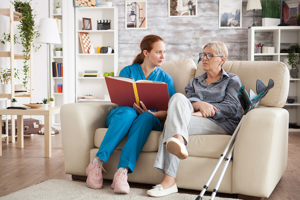 A senior caregiver from Seagrape Home Health Care sitting on a couch and reading to her elderly patient in her home.