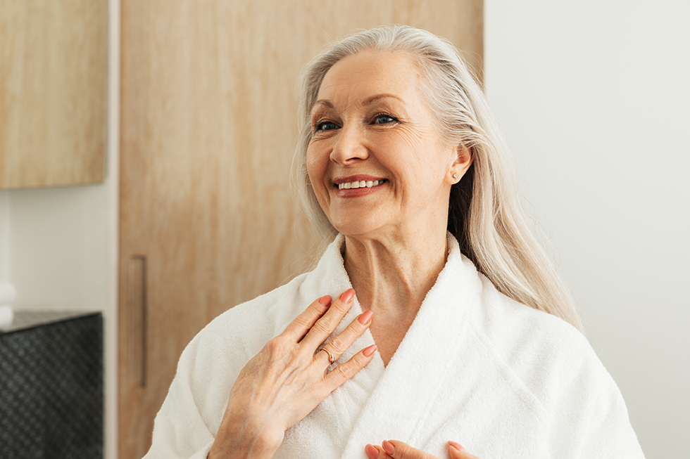 Female senior in her Delray Beach bathroom wearing a white bathrobe standing and smiling | Bathing and Hygiene Caregiver Delray Beach