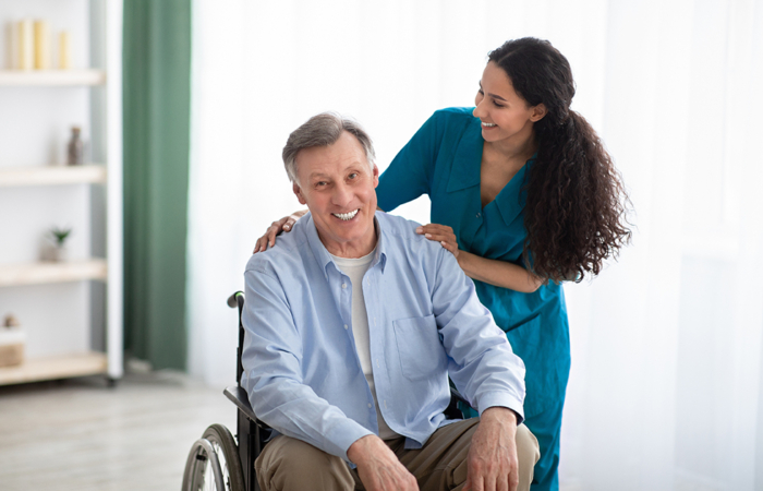 Portrait of happy impaired elderly man and his female nurse at retirement home. Mature handicapped male with his caregiver posing and smiling indoors. Professional medical care for seniors